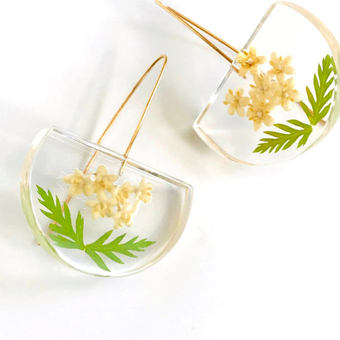 Real elderberry flower earrings. Image shows earrings in a half circle shape with long gold earwires attached in a hook shape. Each earring features six tiny cream colored elderberry flowers and two tiny green leaves floating in a clear eco-resin half moon shape. Eco-resin comes from a tree - not plastic.